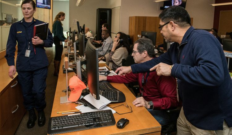 A busy office with people working at computer stations, some seated and others standing while reviewing information on screens.