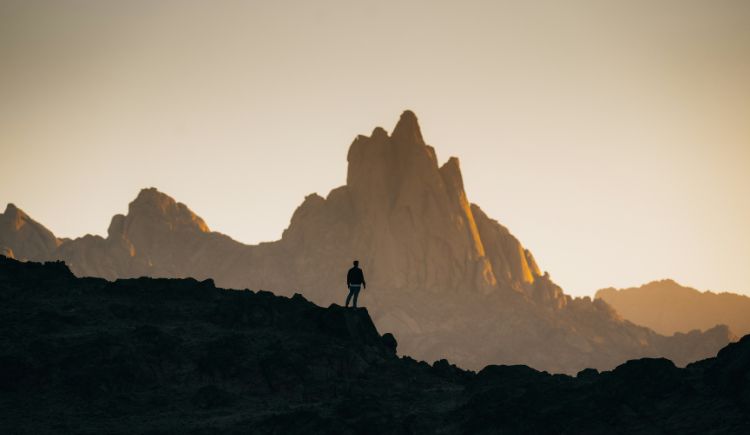 A lone person standing on a rocky ridge, silhouetted against a dramatic mountain range glowing with warm sunrise light.