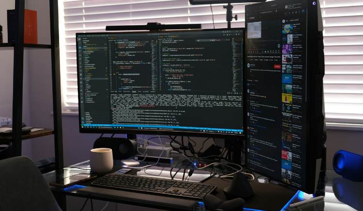 Dual‑monitor desk setup with code displayed on a widescreen monitor and a vertical monitor showing a media feed, along with a keyboard, mouse, and coffee mug on a glass desk.