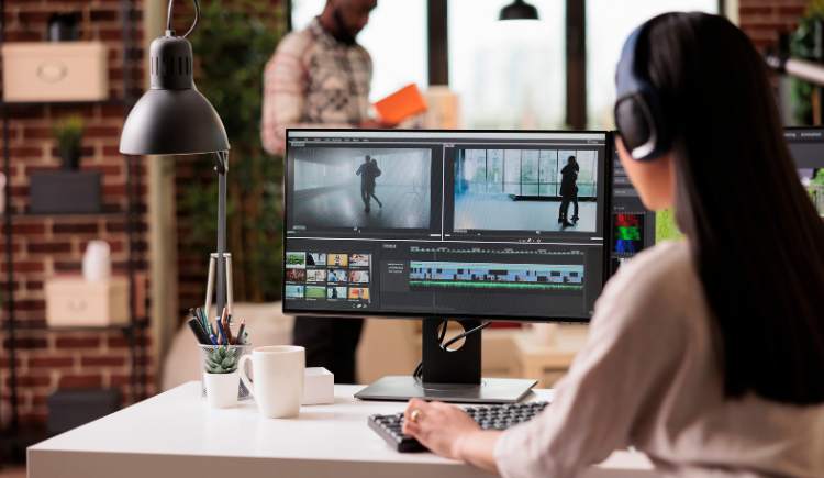 Person editing video on dual monitors at a desk, with another person standing in the background.