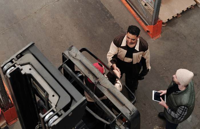 Three warehouse workers gathered around a forklift, with one using a tablet and another interacting with the operator.