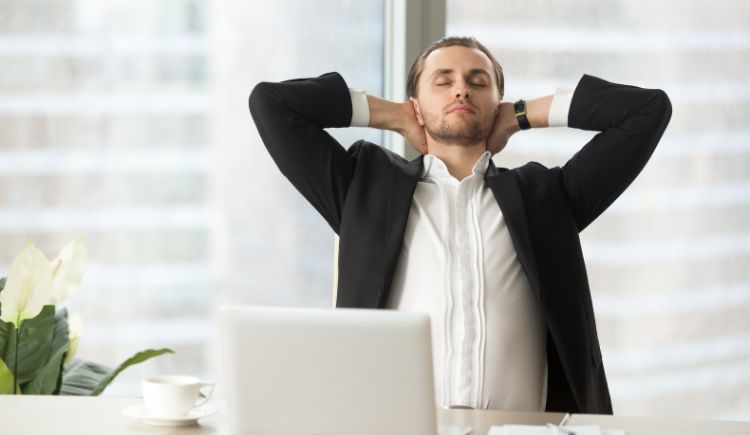 Person in business attire leaning back at a desk with a laptop and coffee cup.