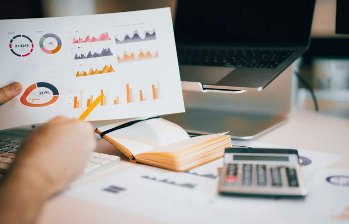 Person pointing to a printed business analytics report with charts and graphs on a desk, next to a laptop, notebook, and calculator.