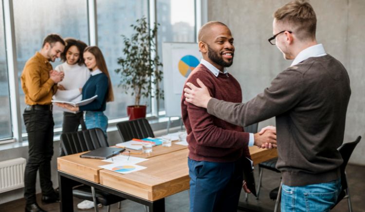 Two coworkers shaking hands in an office while others review documents in the background.