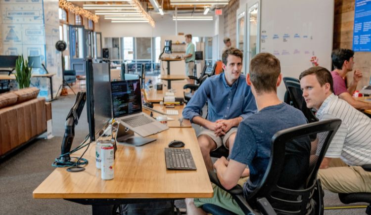 Coworkers collaborating at desks in an open office with computers and shared workspace.